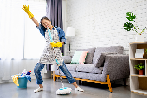 Happy young Asian woman cleaning her home, singing at mop like at microphone and having fun, free space.