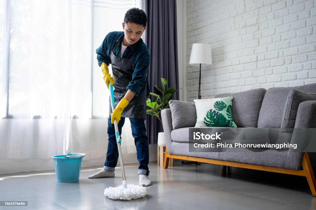 Handsome asian man wearing apron cleaning floor at home. Guy washing floor with mopping stick and bucket in living room. Cleaning Stock Photo