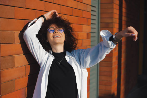 istock Woman posing on brick wall background with curly hair wearing eyewear, closing her eyes with pleasure while smiling in the sunset light. 1363376708