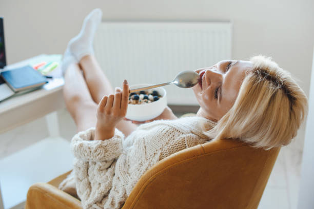 vista para trás de uma mulher branca comendo saboroso café de aveia doce em casa sentado relaxado na poltrona. alimentos orgânicos naturais. - aveia alimento - fotografias e filmes do acervo