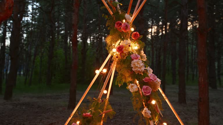 Bohemian tipi wooden arch decorated with burning candles, roses and pampass grass, wrapped in fairy lights illumination on outdoor wedding ceremony venue in pine forest at night. Bulbs garland shines