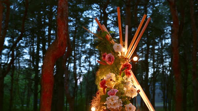 Bohemian tipi wooden arch decorated with burning candles, roses and pampass grass, wrapped in fairy lights illumination on outdoor wedding ceremony venue in pine forest at night. Bulbs garland shines