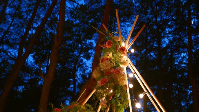 Bohemian tipi wooden arch decorated with burning candles, roses and pampass grass, wrapped in fairy lights illumination on outdoor wedding ceremony venue in pine forest at night. Bulbs garland shines