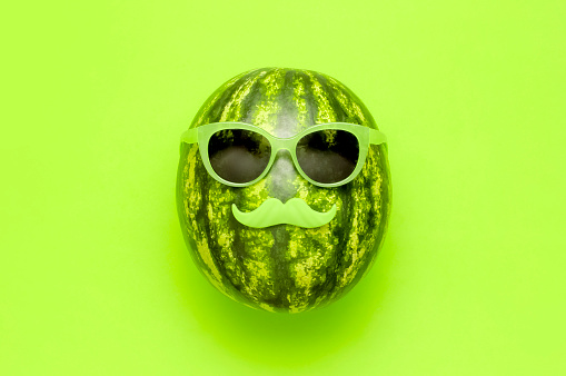Summer funny watermelon in green glasses and with green mustache on bright green background top view flat lay. Minimal fruit concept. Creative product idea, summer festival, summer background.