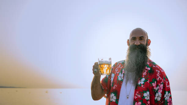 bezwłosy mężczyzna z długą brodą w hawajskiej koszuli trzymający szklankę piwa - beer glass mustache beer color image zdjęcia i obrazy z banku zdjęć