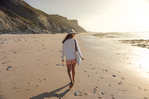 photo of young woman wearing a hat walking along the shore of the beach in a beautiful sunset in Portugal