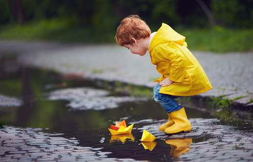 cute little baby boy launching paper boats in spring puddles, wearing raincoat and rubber boots
