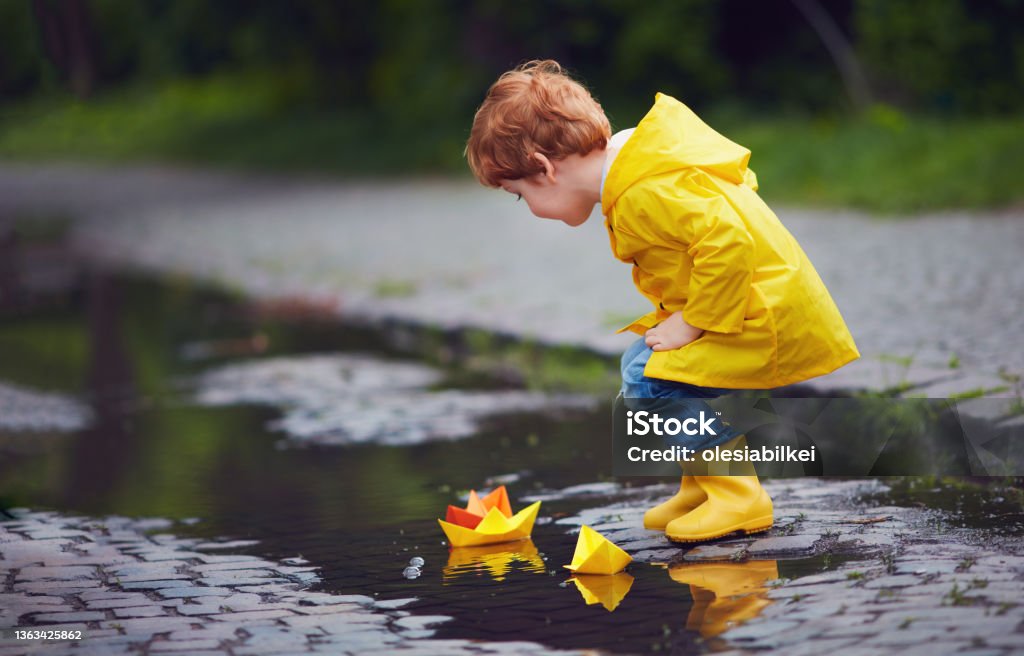 cute little baby boy launching paper boats in spring puddles, wearing raincoat and rubber boots Rubber Boot Stock Photo
