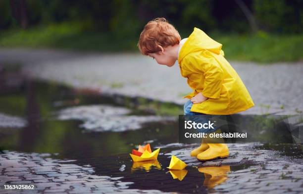 Cute Little Baby Boy Launching Paper Boats In Spring Puddles Wearing Raincoat And Rubber Boots Stock Photo - Download Image Now