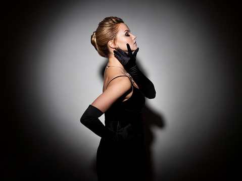 A femme fatale in a black dress with an open back and long velvet gloves. Attractive young blonde with makeup for a celebration, portrait in a spot of light