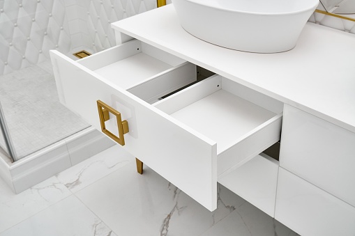 Elegant white cabinet with open drawer and shiny golden handles and leg with vessel sink in contemporary bathroom closeup