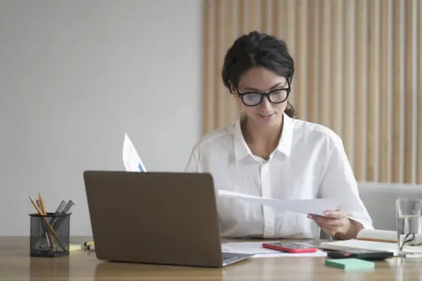 Young focused Italian businesswoman in formal wear analyzing documents, reading project statistics while working on laptop at her workplace, female employee in glasses preparing financial report