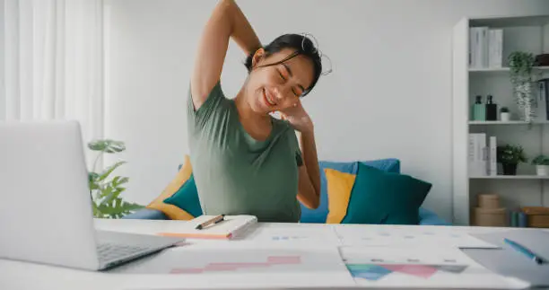 Photo of Beautiful young Asian business woman stretching body for relaxing while working on laptop in living room at home.