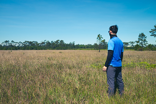Young man standing in a deserted meadow He was looking out into the vast grassland. with a background of sky and pine trees., Rear view , man portrait on landscape , Phu Kradueng National Park ,Thailand
