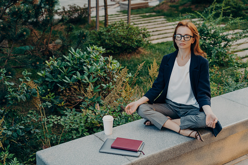 Peaceful calm redhead business woman doing yoga with closed eyes outdoor, relaxing in lotus pose during remote work in park, female relieving stress while working on laptop outside. Meditation concept