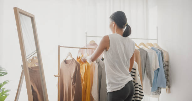 hermosa joven asiática eligiendo ropa en perchero mirándose en el espejo en la sala de estar de casa. - placard women holding standing fotografías e imágenes de stock