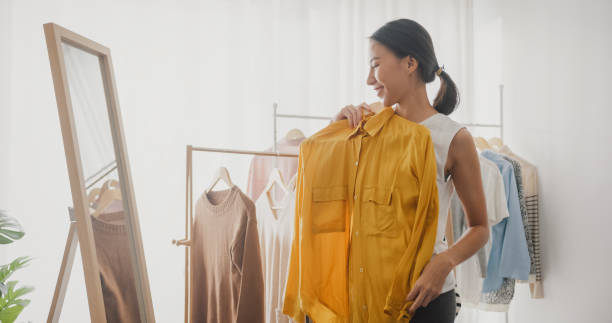 schöne junge asiatische frau, die kleidung auf kleiderständern auswählt, die sich im spiegel im wohnzimmer zu hause ansieht. - women choosing choice clothing stock-fotos und bilder