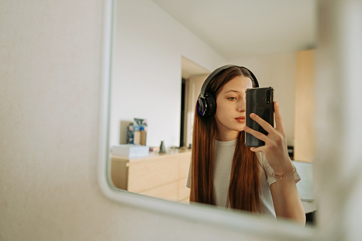 A beautiful girl teenage with long hair ,generation Z in headphones at the mirror in room takes selfie on smartphone, admires herself. Home life, lifestyle. Selective focus