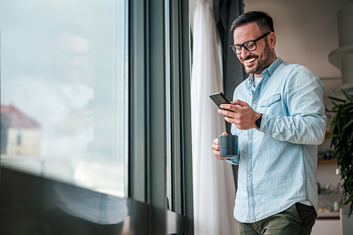 Smiling man businessman in casuals standing in office next to window using mobile phone drinking coffee holding coffee cup Small business entrepreneur looking at smart phone while taking coffee break