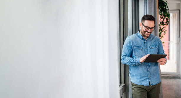 Smiling man businessman in casuals standing in office next to window using tablet making online shopping purchasing Small business home office entrepreneur looking at tablet making online video call