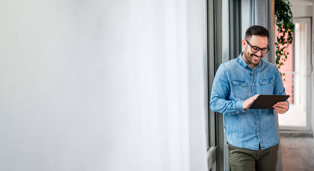 homem sorridente empresário em casuals parados no escritório ao lado da janela usando tablet fazendo compras on-line pequenas empresas home office empreendedor olhando tablet fazendo chamada de vídeo on-line - businessman happiness carefree computer - fotografias e filmes do acervo