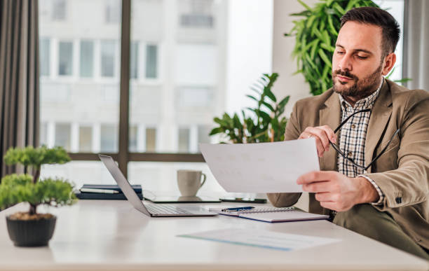 sérieux pensif réfléchi réfléchi axé jeune adulte occasionnel comptable d’entreprise comptable au bureau travaillant avec un ordinateur portable et des documents de déclaration de revenus et des documents en regardant les papiers graphiques graphi - spreadsheet computer report laptop photos et images de collection