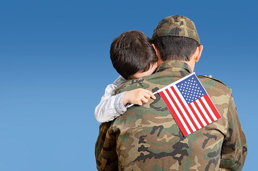 Shot of an adorable child hugging his father affectionately with the united states flag in his hand with a blue sky and copy space.
