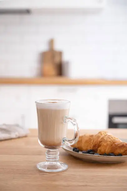Glass of fragrant cappuccino coffee with cinnamon and croissant on a wooden  table against the background of a white kitchen in the early morning. Breakfast concept. Front   "nview.