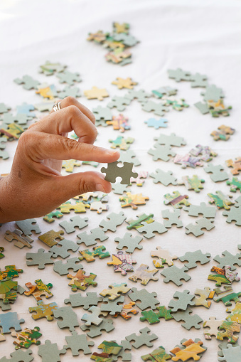Hand of an unrecognizable senior woman is alone playing puzzle on white table.
