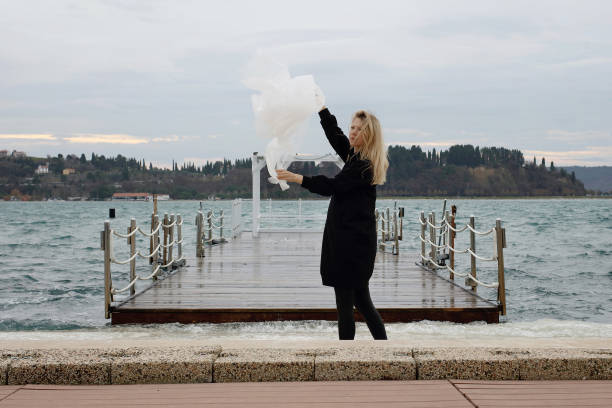 donna vicino al mare nella ventosa giornata invernale che tiene un lungo tessuto che soffia nel vento. concetto d'amore. ballando con la tempesta - women courage water floating on water foto e immagini stock