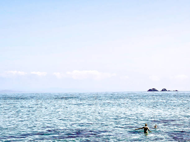Horizontal seascape of two people swimming in the rippled blue sea on a sunny day at Wategos Beach, Byron Bay. Popular scuba diving place, Julian Rocks, in the Cape Byron Marine Park, is visible on the far horizon.