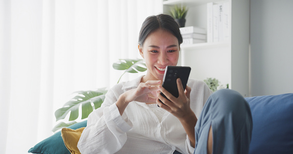 Asian woman using smartphone while sitting on sofa in living room at home.