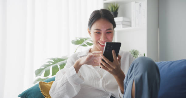 young asian woman using smartphone while sitting on sofa in living room at home. - lying down women laptop freedom imagens e fotografias de stock