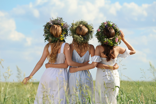 Young women wearing wreaths made of beautiful flowers in field on sunny day, back view