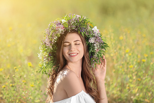 Young woman wearing wreath made of beautiful flowers in field on sunny day