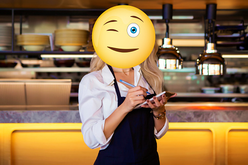 The photo mainly consists of a blonde and pretty woman on whose head an emoji with a blinking eye was put. She is working in a stylish cafeteria in which there is pretty dim light. Classic and nice chandeliers are also put in this place. In the background, there are yellow and brown chairs that look rather comfortable and enticing to sit on. The woman is waiting for the orders of the customers before she begins to service them. This is a workplace, working, coffee house, chilling concept.