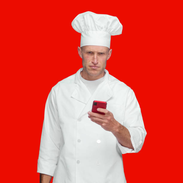 istock Caucasian male chef standing in front of colored background wearing jacket and using text messaging 1363503923