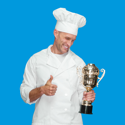 Front view of with short hair caucasian male chef standing in front of blue background wearing pants who is successful and winning and showing award who is in first place and holding trophy