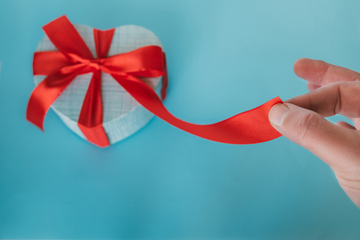 Male fingers holding red ribbon leading to gift. Focus on fingers. Gift box with a bow.