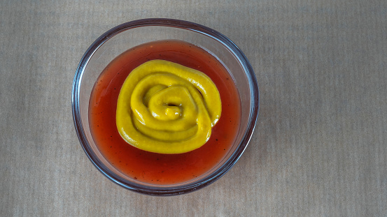 Ketchup with mustard in one glass saucepan on a parchment background. Tasty and choice concepts.