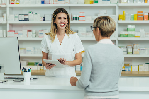 Female pharmacist selling medications at drugstore to a senior woman customer while using tablet