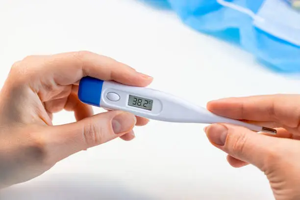 Woman hands holding a medicine digital thermometer with high body temperature measurement on light background. Fever, flu illness and coronovirus desease symptoms concept.
