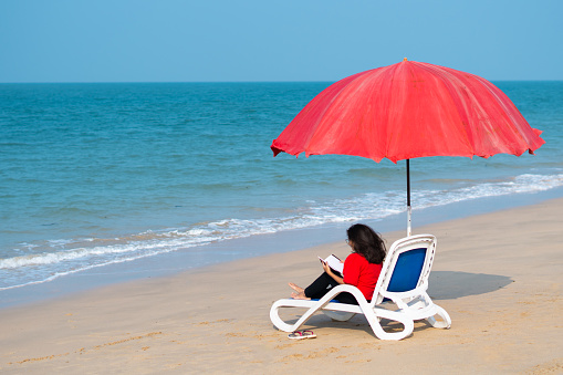 Portrait of young Indian woman sitting on chair under umbrella at tropical beach and reading book. Concept of travel and leisure activities
