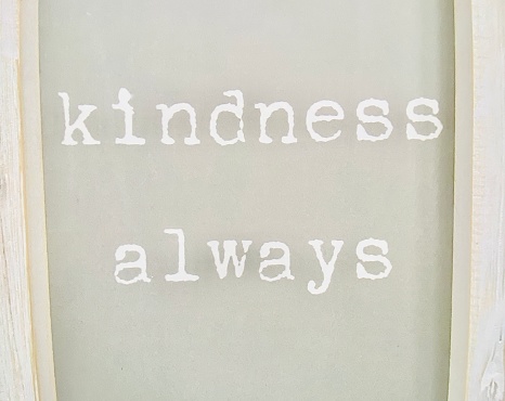 Horizontal sign in neutral tones on domestic kitchen chalk board of text with typewriter font saying: “Kindness Always” with light wood frame