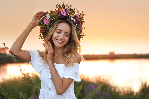 Young woman wearing wreath made of beautiful flowers outdoors at sunset