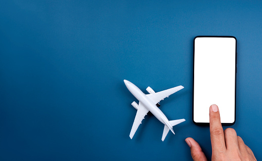 White empty screen on smartphone with touching finger and airplane model toy on blue background with copy space. Air ticket booking on mobile phone app, online travel, and vacation insurance concepts.
