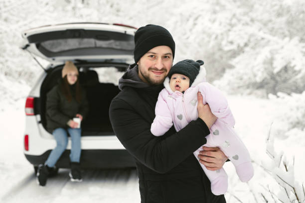 il giovane padre tiene in braccio una bambina sullo sfondo di un'auto in una foresta invernale coperta di neve. - snow bound foto e immagini stock