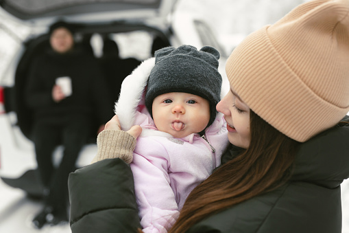 Baby girl looks into the camera and shows her tongue in her mother's arms in a winter snow-covered forest.