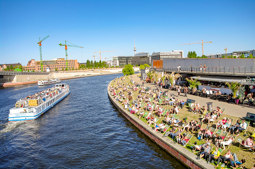 View over relaxing people at the cafe Capital Beach at Spree channel, Spreebogen, with a tour boat in historical and business downtown in Berlin, Germany, at summer sunny and blue sky