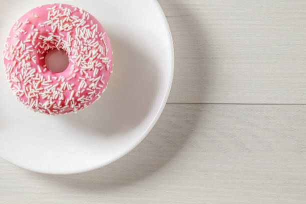 Donut with pink glaze, lying on white plate on light wood countertop. Top view.